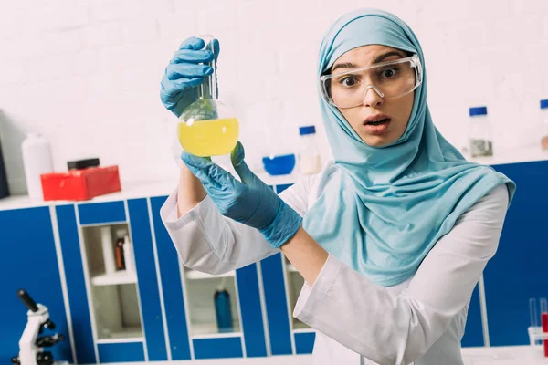 Conmocionado científico musulmán femenino mirando a la cámara y sosteniendo frasco durante el experimento en laboratorio químico - foto de stock