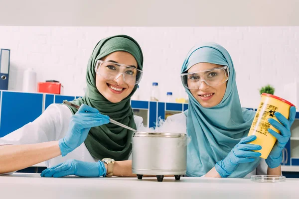 Cientistas muçulmanos fêmeas em óculos olhando para a câmera enquanto experimenta com gelo seco em laboratório químico — Fotografia de Stock