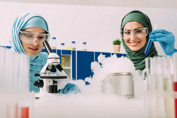 Female muslim scientists in goggles experimenting with microscope and dry ice in chemical laboratory — Stock Photo