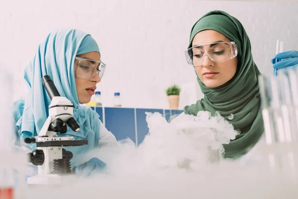 Muslimische Wissenschaftlerinnen in Schutzbrillen experimentieren im Chemielabor mit Mikroskop und Trockeneis — Stockfoto