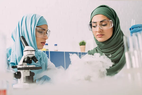 Científicas musulmanas en gafas que experimentan con microscopio y hielo seco en laboratorio químico — Stock Photo