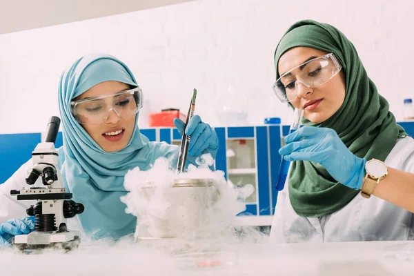 Female muslim scientists in hijab experimenting with microscope and dry ice in chemical laboratory — Stock Photo