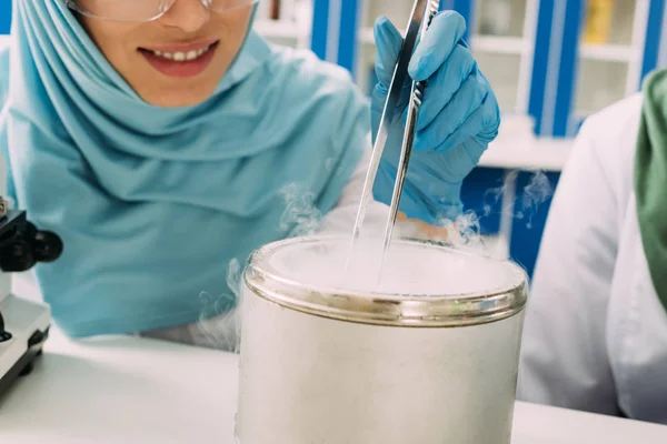 Vie parcial de cientista muçulmano fêmea segurando pinças durante o experimento com gelo seco em laboratório — Fotografia de Stock