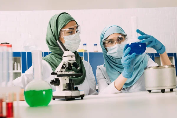 Científicos musulmanes femeninos con frascos y microscopio durante el experimento en laboratorio químico - foto de stock