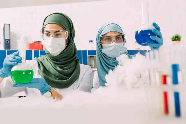 Científicas musulmanas sosteniendo frascos mientras experimentan con hielo seco en laboratorio químico - foto de stock