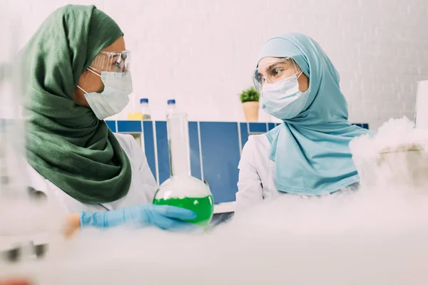 Científicas musulmanas con matraz mirándose mientras experimentan con hielo seco en laboratorio químico — Stock Photo