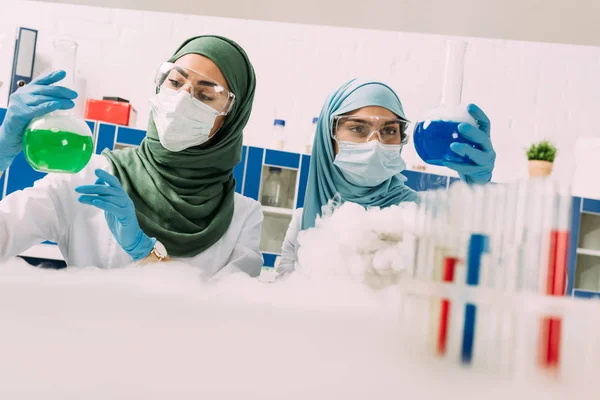 Científicas musulmanas en máscaras médicas sosteniendo frascos mientras experimentan con hielo seco en laboratorio químico — Stock Photo