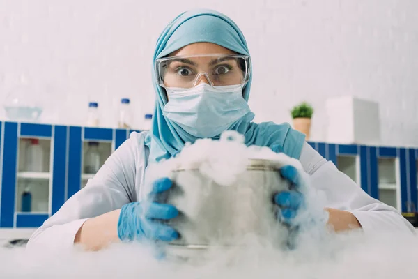 Surpris femme musulmane scientifique regardant la caméra tout en tenant le pot avec de la glace sèche en laboratoire — Photo de stock