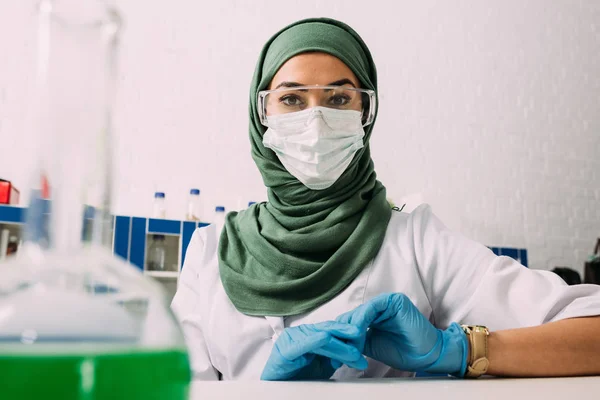 Femme musulmane scientifique en lunettes et masque médical regardant caméra en laboratoire chimique avec fiole au premier plan — Photo de stock