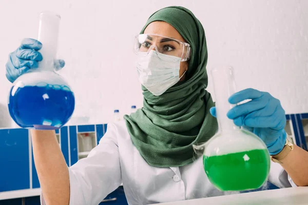 Cientista muçulmano fêmea em máscara médica segurando frascos durante experimento em laboratório químico — Fotografia de Stock