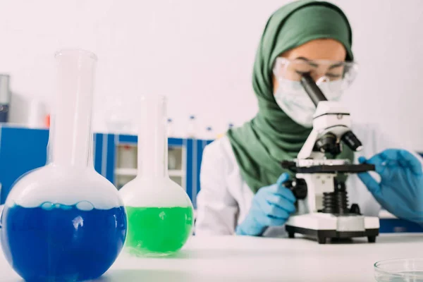Cientista muçulmano fêmea sentada à mesa com frascos e usando microscópio durante o experimento em laboratório químico — Fotografia de Stock