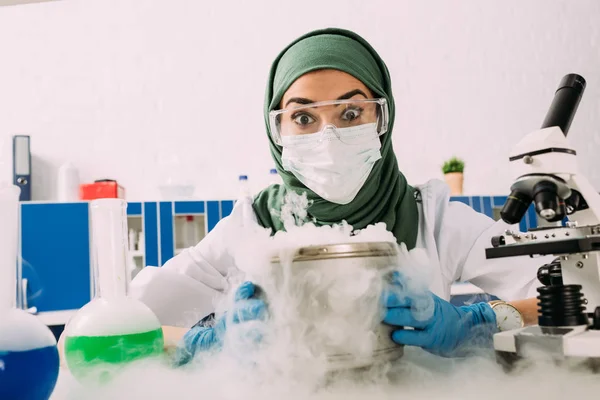 Conmocionado científico musulmán femenino sosteniendo olla con hielo seco durante el experimento en laboratorio - foto de stock