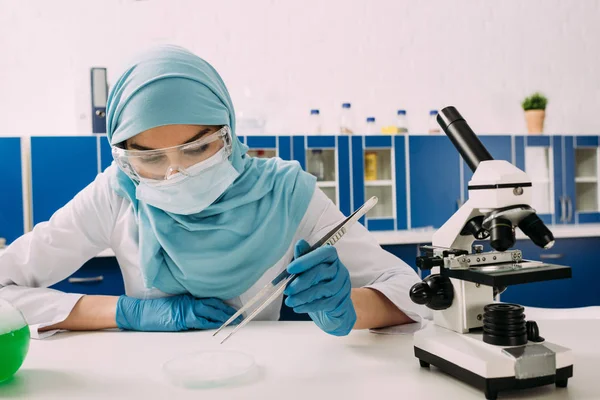 Muslimische Wissenschaftlerin sitzt mit Mikroskop am Tisch und benutzt Pinzette mit Petrischale bei Experimenten im Chemielabor — Stockfoto