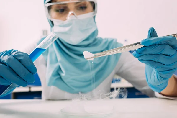 Vista recortada del científico musulmán femenino sosteniendo tubo de ensayo con pinzas y experimentando con hielo seco en laboratorio — Stock Photo