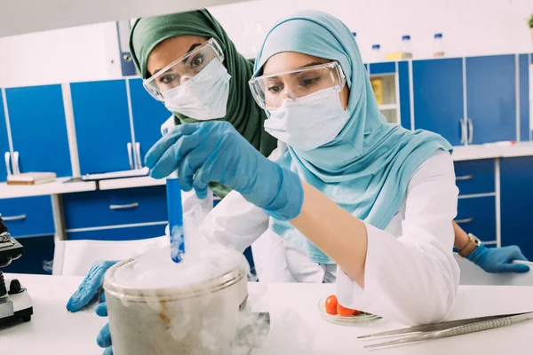 Female muslim scientists in goggles holding reagent over dry ice during experiment in chemical lab — Stock Photo