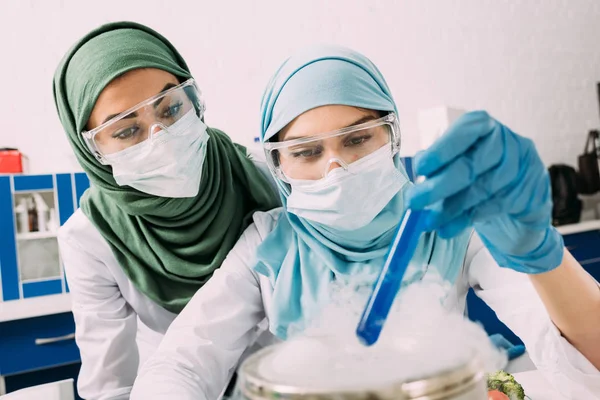 Cientistas muçulmanas fêmeas em óculos segurando reagente sobre gelo seco durante experimento em laboratório químico — Fotografia de Stock