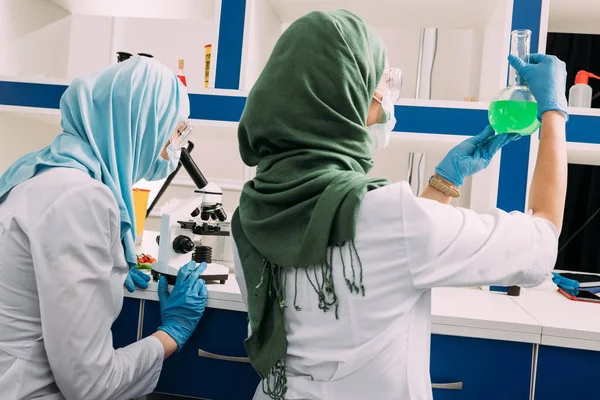 Назад взгляд женщин-мусульманских ученых, держащих фляжку и использующих микроскоп во время экспериментов в химической лаборатории — стоковое фото