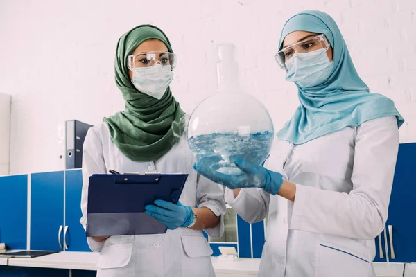 Female muslim scientists holding clipboard and flask wth liquid in chemical laboratory — Stock Photo