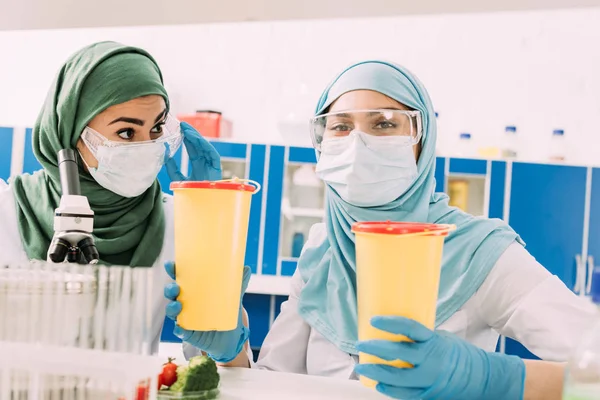 Muslimische Wissenschaftlerinnen halten klinische Abfallbehälter in die Höhe und blicken in das Chemielabor — Stockfoto
