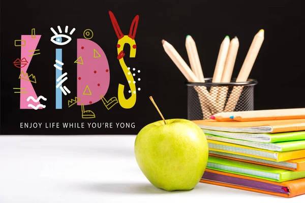 Fresh apple, notebooks and color pencils on table with enjoy life while you are young and kids lettering on black — Stock Photo