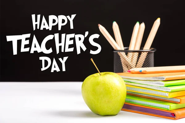 Свежее яблоко, блокноты и цветные карандаши на столе с счастливыми учителями день написания на черном — стоковое фото