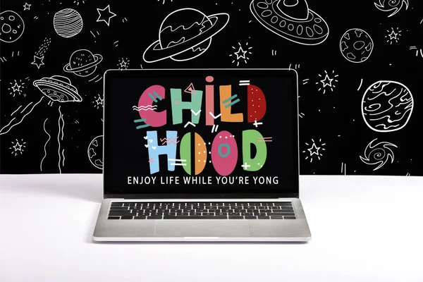 Portátil en la mesa con disfrutar de la vida mientras usted es joven y letras de la infancia en la pantalla con la ilustración del espacio en blanco en negro - foto de stock