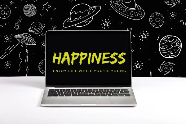 Portátil en la mesa con disfrutar de la vida mientras eres joven y letras de felicidad en la pantalla con la ilustración de galaxia blanca en negro - foto de stock