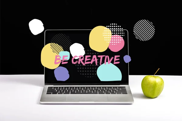 Fresh green apple and laptop on table with be creative lettering on screen and abstract circles around on black — Stock Photo