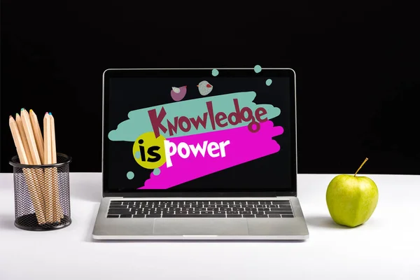 Fresh green apple, color pencils and laptop on table with knowledge is power lettering on screen isolated on black — Stock Photo