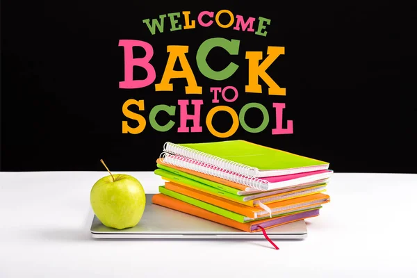 Portátil cerrado, manzana verde, cuadernos y lápices de color en el escritorio con bienvenida de nuevo a las letras de la escuela en negro - foto de stock