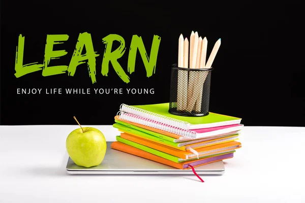 Closed laptop, green apple, notebooks and color pencils on desk with learn lettering on black — Stock Photo