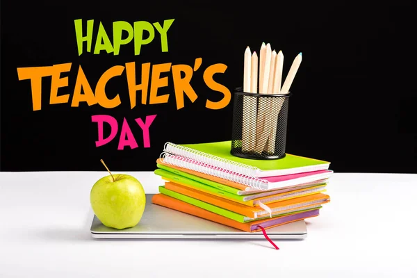 Portátil cerrado, manzana verde, cuadernos y lápices de color en el escritorio con letras felices maestros día en negro - foto de stock