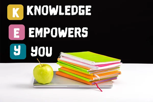 Closed laptop, green apple, notebooks and color pencils on desk with knowledge empowers you lettering on black — Stock Photo