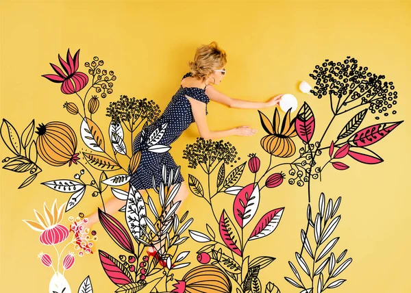 Top view of young elegant woman with saucer and cup lying on yellow background with floral illustration — Stock Photo