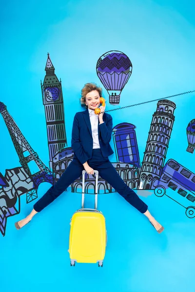 Vista superior de la mujer de negocios en traje con bolsa de viaje hablando por teléfono mientras está acostado sobre fondo azul con ilustración de la ciudad - foto de stock