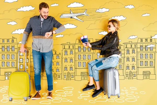 Щаслива жінка тримає паспорти та квитки, а чоловік з валізою перевіряє час з ілюстрацією міста та літака на жовтому тлі — стокове фото