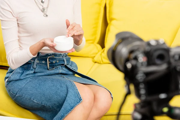 Vista cortada do blogueiro em saia jeans segurando creme cosmético na frente da câmera de vídeo — Fotografia de Stock