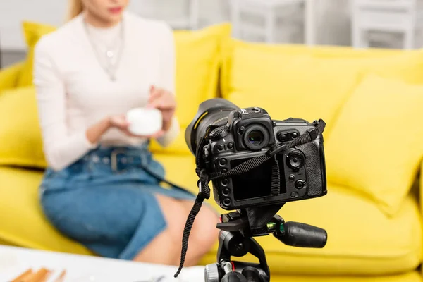 Vista cortada do blogueiro em saia jeans segurando creme cosmético na frente da câmera de vídeo — Fotografia de Stock