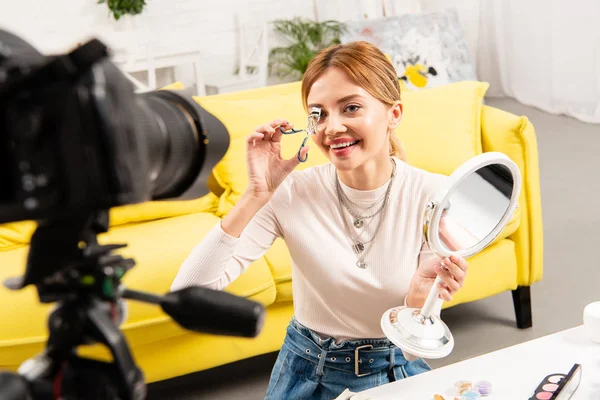 Sonriente belleza blogger sosteniendo espejo y rizando pestañas delante de la cámara de vídeo - foto de stock