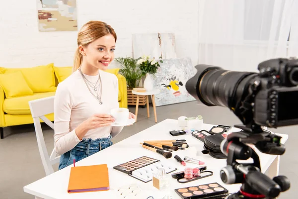 Blogueur beauté assis à table avec des cosmétiques décoratifs et tenant une tasse de café devant la caméra vidéo — Photo de stock