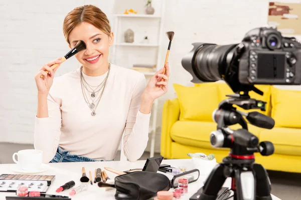 Sonriente blogger de belleza sosteniendo pinceles cosméticos frente a la cámara de video - foto de stock