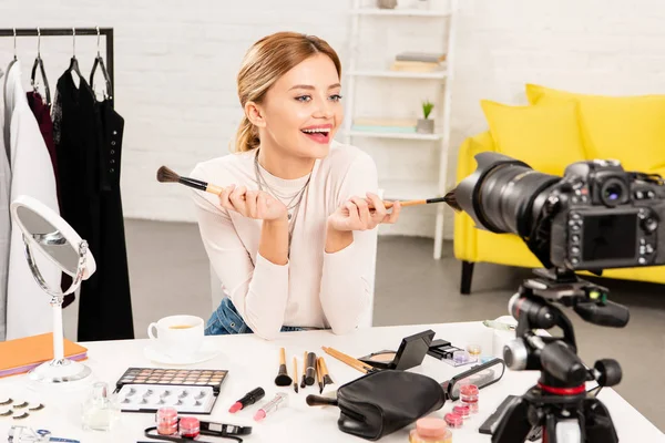 Sonriente blogger de belleza sosteniendo pinceles cosméticos frente a la cámara de video - foto de stock