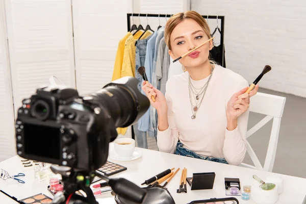 Blogueur beauté avec des cosmétiques décoratifs tenant pinceaux cosmétiques devant la caméra vidéo — Photo de stock