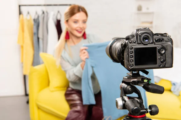 Selective focus of smiling blogger sitting on sofa and showing clothes in front of video camera — Stock Photo