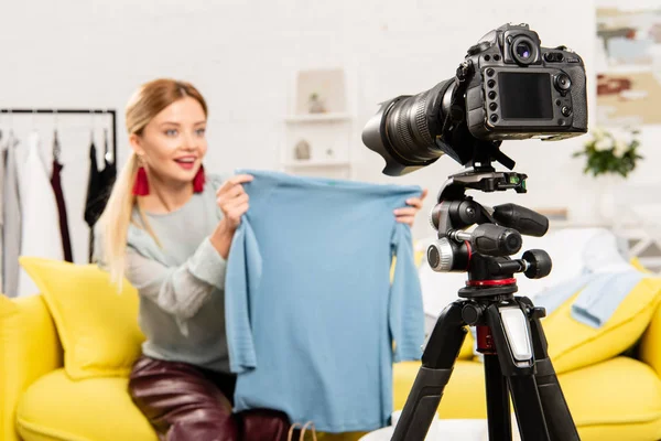 Избирательный фокус улыбающегося блогера, сидящего на диване и показывающего одежду перед видеокамерой — стоковое фото