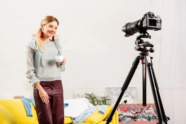 Blogger sonriente en pendientes sosteniendo una taza de café frente a una cámara de video en casa - foto de stock