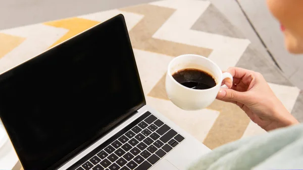 Vista recortada del freelancer sosteniendo taza de café y utilizando el ordenador portátil con pantalla en blanco - foto de stock