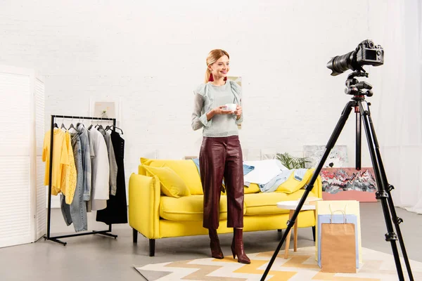 Full length view of smiling blogger holding cup of coffee in front of video camera at home — Stock Photo