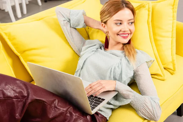 Freelancer sorrindo deitado no sofá amarelo e usando laptop em casa — Fotografia de Stock