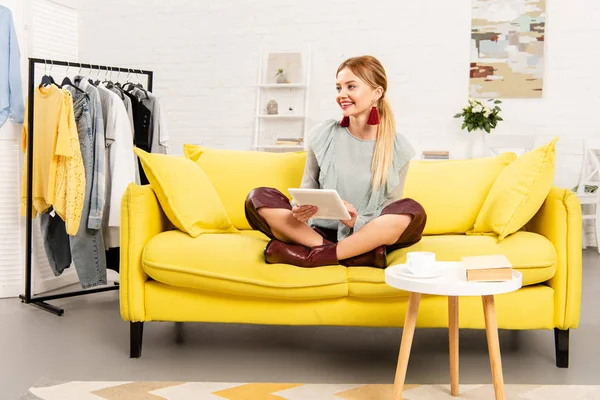 Smiling girl sitting on yellow sofa with crossed legs and using digital tablet — Stock Photo